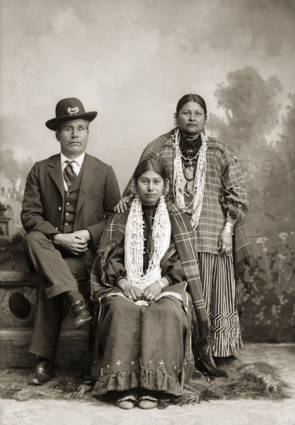 Full-length studio portrait in front of a painted backdrop of a European American man, and two Ho-Chunk women. The man is wearing a military hat and suit jacket, vest, and tie, and the women are wearing long necklaces and ear bobs. Identified as non-Ho-Chunk Charlie Saunders with his Ho-Chunk wife  Mary Eagle (ChaUkShePaRooEWinKah) and her daughter, Annie Davis Bigsoldier White Rogue (KeeReeJayWinKah). Saunders served in the Civil War as a private in Company B of the 1st Wisconsin Cavalry and was twice a prisoner of war. The first time, he was captured in Bloomfield, Missouri, and released in exchange for a Confederate prisoner. He was captured again on July 30, 1864, and held at the infamous Andersonville Prison. Saunders is wearing the GAR (Grand Army of the Republic) emblem on his hat. Saunders spoke the Ho-Chunk language and was close to the Ho-Chunk community, but it is not known whether he received Ho-Chunk warrior privileges.