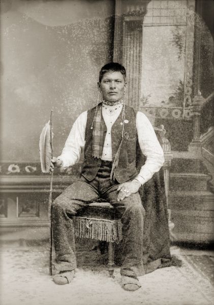 Studio full-length portrait of a Ho-Chunk man with short hair posing sitting in front of a painted backdrop. Identified as John Johnson. John was a private with Company A of the Omaha Scouts. He is wearing traditional Ho-Chunk moccasins and earrings. In his hand is a staff with an eagle feather attached, indicating his warrior status.