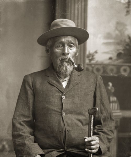 Waist-up studio portrait of a Ho-Chunk man with a beard posing sitting in front of a painted backdrop. He is smoking a pipe and holding a cane in his left hand, and is wearing a suit and hat. He is identified as John Hazen Hill (XeTeNiShaRaKah). He served in the Civil War with the 14th Kansas Volunteer Cavalry Regiment and was reportedly of mixed blood, with a European American father.