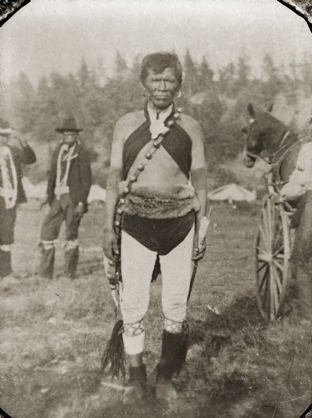 A Ho-Chunk man is posing standing in a field, holding an object in his left hand, and is wearing regalia including a long bandoleer which appear to have bells attached, light-colored leggings, a fur belt, and dark breech cloth. He is identified as George Standingwater (HaNaKah), an army veteran. He was of the Blue Wing Band, clan name Water Spirit Little Ocean Waves. In the background there are two men standing on the left, and a horse pulling a wagon or carriage on the right. Tents are in the far background.