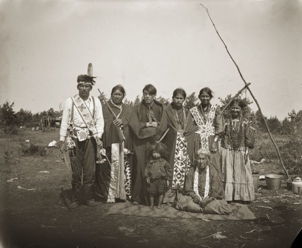 A Ho-Chunk group dressed in regalia standing behind an elderly woman, sitting, and child who is standing. The man with the rifle has an appliqué blanket wrapped around him, as does the woman in the middle of the back row, third from left.