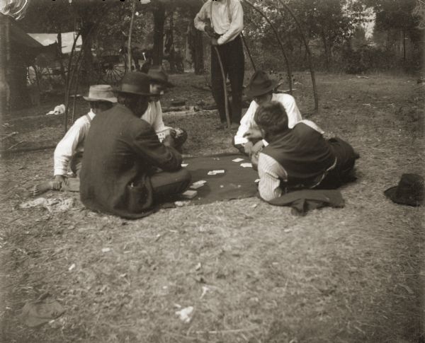 Five Ho-Chunk men are sitting around a blanket on the ground playing cards. They are in front of a lodge frame without coverings. A man holding a walking stick is standing behind them watching. There are horses and a wagon in background.