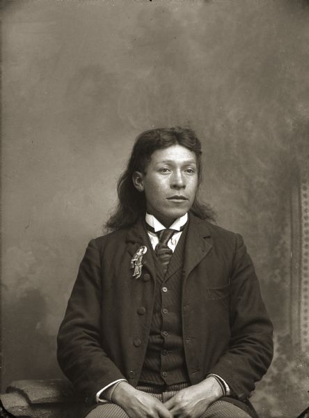 Waist-up studio portrait in front of a painted backdrop of a Ho-Chunk man with long hair posing sitting. He is wearing a suit, necktie, and a button with a photograph picturing a man with long hair. Identified as Henry Greencrow (CooNooZeeKah). Henry has a button on his lapel made from a photograph. Some of Van Schaick’s photographs were made into buttons like this one.
