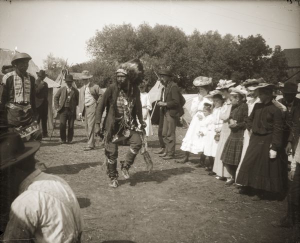 An unidentified man dancing for a group of onlookers in Black River Falls during the 1908 Homecoming Celebration. He is wearing a Sioux-style eagle feather headdress and is carrying a Plains-style pipe bag.