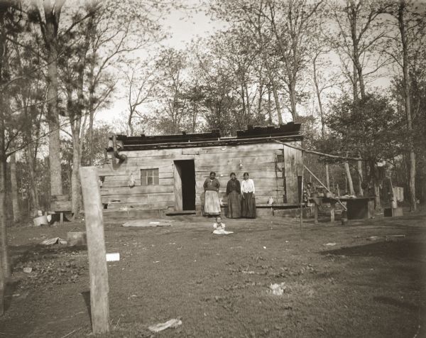 View across yard towards three unidentified women and an infant posing in front of a log home located near Wittenberg. It seemed strange to many Ho-Chunk to live in a house that was square. Very few Ho-Chunks lived in log or framed houses until the WPA built housing for the Ho-Chunk.