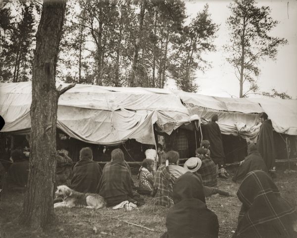 Unidentified Ho-Chunk women sitting outside of a Medicine Lodge, whose sides are rolled up to allow airflow. Only members of the Medicine Lodge were allowed to enter the lodge and participate in the ceremonies.