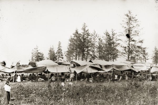 A Medicine Dance Lodge at Morrison Creek. There is a long tent in a field with a large group of people and children sitting and standing underneath it. There is also a wagon in front of the tent on the far right side. Trees are in the background, and in the foreground is a child standing on the left.