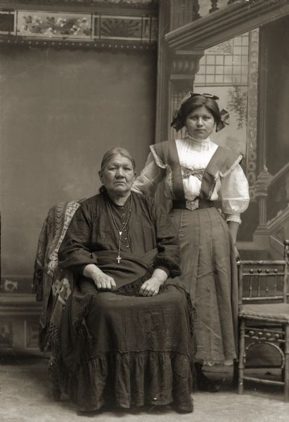 Full-length atudio portrait in front of a painted backdrop of Lucy Goodvillage Blowsnake (NauNawZokeAWinKah) sitting beside her granddaughter Nina Thunder Decorah Green (AhHooGeNaWinKah), who is standing on the right. Lucy is wearing a Christian cross around her neck.