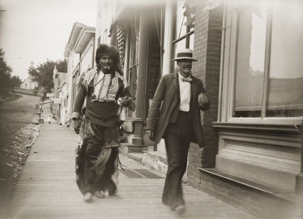 View down wooden sidewalk towards a Ho-Chunk man smoking a cigar and wearing a duck bone breast plate and fur pants on the left, and a European American man wearing a suit, bow tie, and straw hat on the right, along Main Street, prior to the 1911 flood. There is a church building in the far distance. There is a child standing on the sidewalk in the far background.