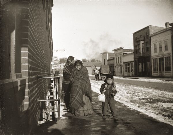 Two Ho-Chunk girls wrapped in Racine Woolen Mills shawls walking with a young boy down Water Street downtown. The Journal sign is visible behind them, and there is a horse-drawn carriage on the street in the background.
