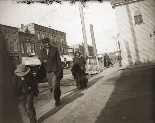 Jim Swallow (MaPaZoeRayKeKah) and Pinkey Bigsolder (HoWaWinKah), as well as an unidentified child, crossing at the corner of Main and South First Street. Jim Swallow was a tall man, at least six and a half feet in height, who lived between Sandy Plains and Shamrock. Early explorers who had contact with the Ho-Chunk often noted how tall the Ho-Chunk were. A poster in the background advertises a show at the Opera House on Wednesday, May 6.