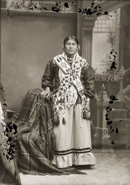 Full-length studio portrait in front of a painted backdrop of Blanche Blackhawk (Hopinkah) (ENookskaGaKah) standing next to a chair covered in a Racine Woolen Mills shawl (wai). These shawls were marketed exclusively to Native Americans. She is wearing a woman’s dress (hinukwaje) and traditional earrings(naacawa hocpok), rings (naap hirusigic), and bracelets (suura sgaa aipa). She wears a necklace made of long strands of shell and beads and two Ho-Chunk bandolier bags.