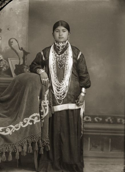 Studio portrait of a young Ho-Chunk woman posing standing in front of a painted backdrop. Her right arm is resting on a draped chair and she is wearing several necklaces, earrings, rings, and file bracelets. Identified as Lucy Dot Redbird Brown (WakJaHeWinKah).