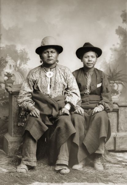 Two unidentified Ho-Chunk men posing in front of a painted backdrop. The man on the left is wearing earrings, a shell and metal bead necklace, and traditional Ho-Chunk-style moccasins. On the back of the vintage print of this image, one of the men is identified as Thomas Lolly (?).