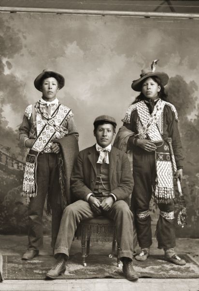 Charlie Greengrass (HoeHumpCheeKayRayHeKah), left, and David Bow Littlesoldier (MauJchayMauNeeKah) ar standing behind Will Stohega Carriman (WonkShiekStoHeGah). Will is dressed in contemporary 1890s clothing, while the others are wearing traditional regalia.