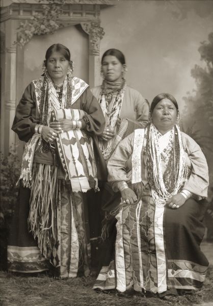 Full-length studio portrait in front of a painted backdrop of three unidentified women. The woman sitting on the right has contrasting color stitching around the floral designs on her appliqué skirt. The woman on the left is wearing two Ho-Chunk beaded bandolier bags and is holding a silk appliqué blanket.