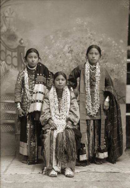 Full-length studio portrait of three young Ho-Chunk woman posing in front of a painted backdrop. Addie Littlesoldier Lewis (KzunchJeKahRayWinKah)is sitting between Queen Redbird Yellowbank, left, and Sally Dora Redbird Goodvillage Whitewater (WauNeekShootchWinKah). The two girls that are standing are cousins; their mothers are sisters.