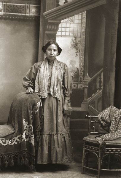 Full-length portrait of Annie Lizzie Falcon Brown Whitedog Hall (DasChuntAWinKah) standing in front of a painted backdrop. She is wearing a traditional Ho-Chunk dress (hinukwaje) and is resting her hand on an embroidered blanket that was a prop in Van Schaick’s studio. This blanket shows up in many photographs throughout Van Schaick’s career.