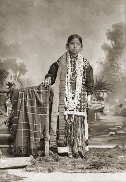 Full-length studio portrait of Sally Dora Redbird Goodvillage Whitewater (WauNeekShootchWinKah). The braided ends of her finger woven sash are hanging down from her waist, covering up the appliqué on her skirt. The appliqué on the skirt was traditionally worn to the side but was sometimes brought to the center so that it could be seen in photographs. She has her hand resting on a blanket draped over a prop wooden fence.