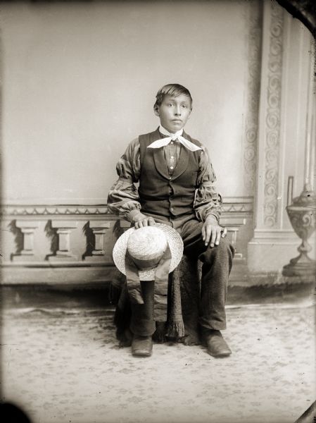 Full-length studio portrait in front of a painted backdrop of an unidentified boy holding a hat with a wide ribbon band on his knee.