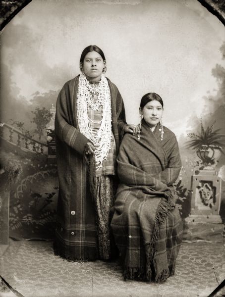 Studio full-length portrait of two unidentified Ho-Chunk women posing in front of a painted backdrop. The woman standing on the left is wearing several necklaces, earrings, and a plaid fringed shawl over her shoulders, and the Ho-Chunk woman posing sitting on the right is wearing long earrings and is wrapped in a plaid fringed shawl.