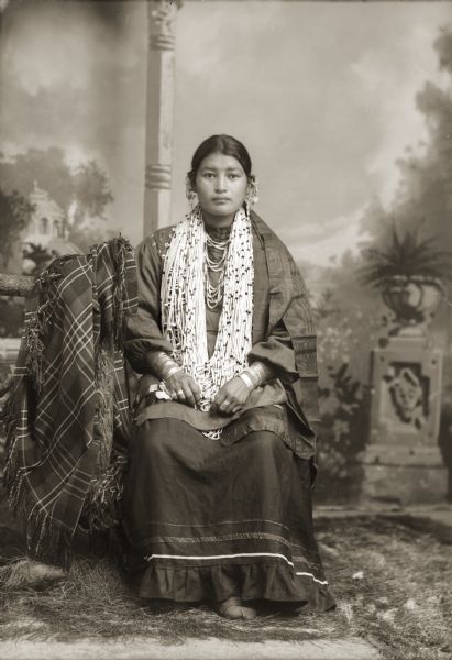 Full-length studio portrait in front of a painted backdrop of a young woman displaying her shell necklace, silver bracelets, and earrings.
