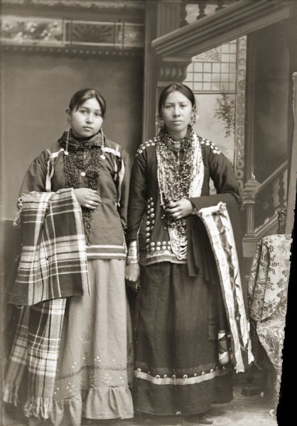 Studio portrait of two Ho-Chunk women posing standing in front of a painted backdrop. They are wearing several necklaces, earrings, and rings. Both have shawls draped over their outside arms, although the women's shawl on the right has ribbon work. Identified as Lena Ora Longthunder (UkSeKaHoNoKah), left, and Alice Thunderking Lowe Kingswan (MaHayPaSayWinKah).