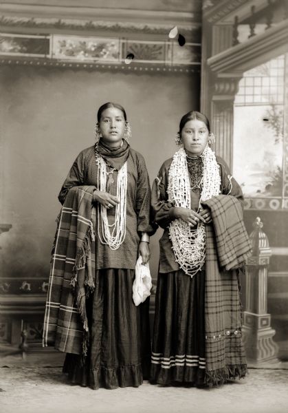 Full-length studio portrait in front of a painted backdrop of two young Ho-Chunk women. They are both wearing several necklaces and earrings, and are each holding a plaid shawl over their outside arm. Identified as Lucy Decorah (AhHoRaPaNeeWinKah), left, and Alice Standstraight Blowsnake Littlewolf (HeChoWinKah).