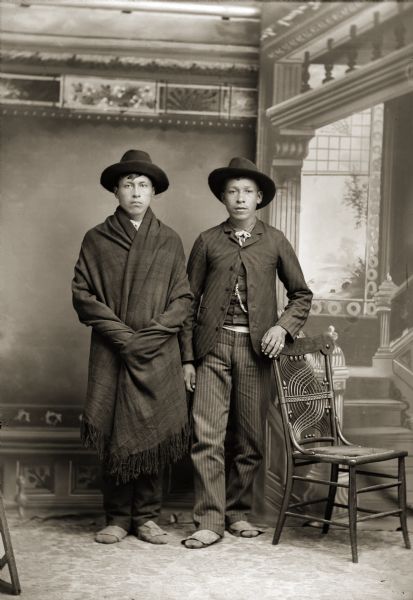 Full-length studio portrait in front of a painted backdrop of two Ho-Chunk men posing standing and wearing hats . The man on the left, John Stacy (ChoNeKayHunKah), has a blanket wrapped around his shoulders. The man on the right, Henry Greencrow (CooNooZeeKah), has his hand resting on the back of a chair. Both men are wearing traditional Ho-Chunk moccasins.