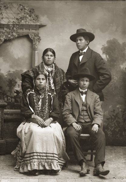Studio portrait in front of a painted backdrop of two Ho-Chunk couples. Albert Henry (HahNahKah) is standing with his wife, Martha Henry. Sitting in front are Martin Green (Snake) (KeeMeeNunkKah) and his wife, Dora Monegar Wallace Green (Snake) (ChePinChayWinKah). The women are wearing several necklaces, earrings, and file bracelets. The men are wearing suits, bow ties, and hats.