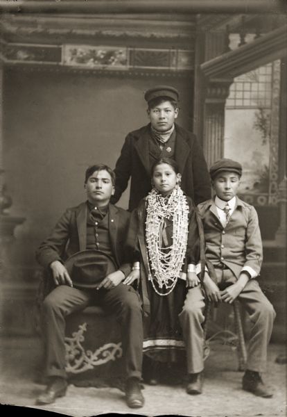 Studio portrait in front of a painted backdrop of a Ho-Chunk man posing standing wearing a winter coat and hat. On the left is a Ho-Chunk man sitting and wearing a suit and holding a hat in his lap. In the center is a Ho-Chunk girl who is standing and wearing a large wampum necklace and earrings. On the right is a Ho-Chunk boy sitting on a stool and wearing a suit, necktie, and cap.
