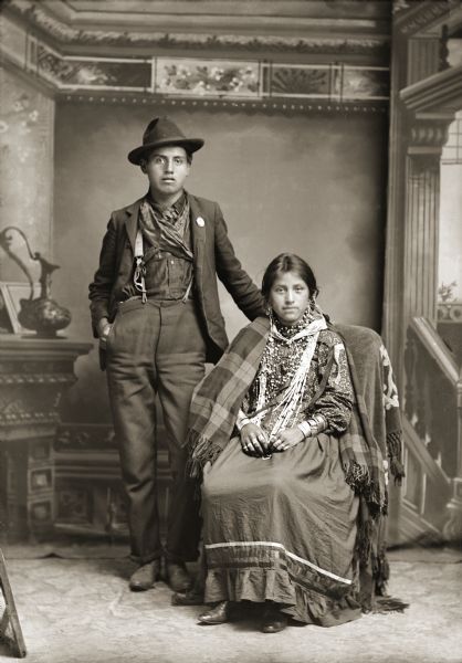 Studio portrait of a Ho-Chunk couple. Johnnie Thunder (Little Chief) (Hoonk Neek Kah) standing on the left and wearing a dark-colored suit and hat, with his right hand behind the right shoulder of his seated wife. Ida Whitespirit, Thunder (Wauk Che He Ska Pe He Ta Win Kah) is wearing several necklaces, earrings, file bracelets, rings, and a shawl draped over her shoulders. In the background is a painted backdrop.