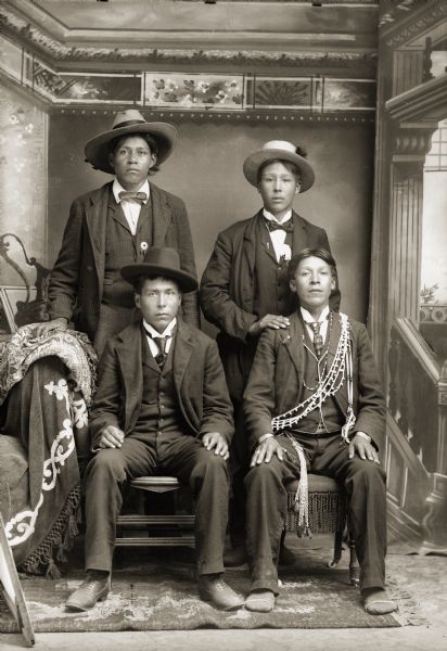 Studio portrait in front of a painted backdrop of two young Ho-Chunk men posing sitting in front of two men posing standing, William Hall (HunkKah), left, and Charlie Greengrass (HoeHumpCheeKayRayHeKah) on the right. The men are wearing contemporary clothing, such as suits, bow ties, and hats. Henry Greencrow (CooNooZeeKah), sitting on the right, is wearing Ho-Chunk moccasins, and a bone-and-shell bandolier over his shoulders.