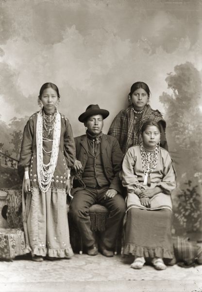 Full-length studio portrait in front of a painted backdrop of a Ho-Chunk man and three Ho-Chunk women. The woman standing on the left is wearing many necklaces, earrings, file bracelets, and a silver drop-fringed blouse. The man sitting in the center is wearing a hat, earrings, a suit coat and jacket, and a bandana. The woman sitting on the right is Grace Decorra Twocrow Winneshiek Massey (MauHeHutTaWinKah), and she is wearing a blouse and necklaces. The woman standing behind her Ida Lizzie Decorah Blowsnake (Real Wampum Woman) (WooRooShiekESkaeWinKah) is wearing necklaces, earrings, and a shawl wrapped around her shoulders. The group is identified as probably the King of Thunder Family according to the notes of Jackson County Historical Society, Wisconsin.