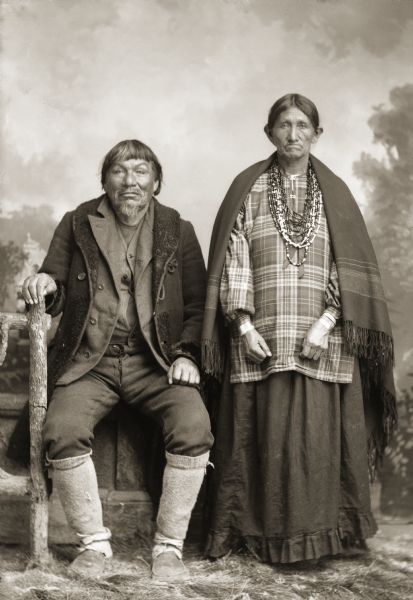 Full-length studio portrait in front of a painted backdrop of a Ho-Chunk couple, Blackhawk (KaRaChooSepMeKah), and his wife Louisa Mike (HumpACooWinKah). Blackhawk is posing sitting on a prop stone wall with his hand resting on a prop wooden fence. He is wearing a coat, suit jacket, vest, trousers, and leggings. Louisa Mike is standing and wearing a dress with a blouse, necklaces, bracelets, and a shawl over her shoulders.