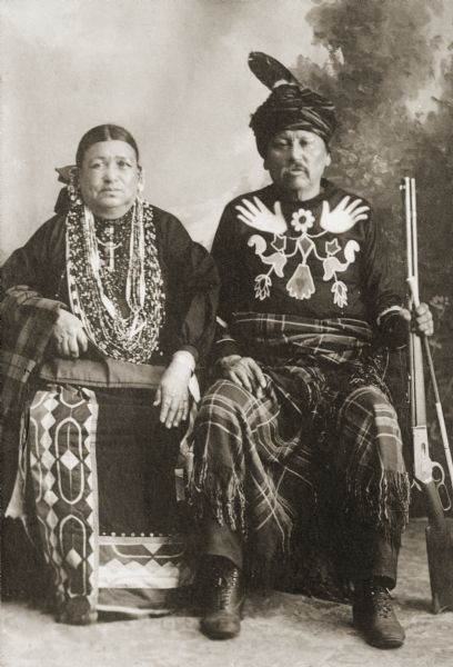Copy photograph of a studio portrait of Ho-Chunk couple, Little Soldier (NoGinKah) and his wife Bettie Littlesoldier (BayBayBawKah)  sitting and wearing regalia in front of a painted backdrop. Little Soldier is holding a rifle. He was also known as "Strike the Tree," according to the notes of Jackson County Historical Society, Wisconsin. According to the Census of the Wisconsin Winnebago Indians in 1905, the Soldier's had a daughter named Ellen.