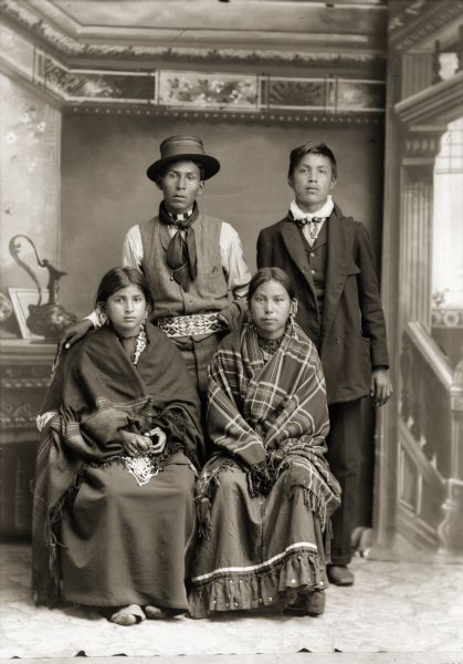 Studio portrait of Ho-Chunk group, Frank Standingwater (WaTaChayHoNeeKah), left, and Frank Redbird (WakJaHeNoKah) standing behind Lucy Dot Redbird (WakJaHeWinKah), left, and Jennie Bighawk-Devorra Mallory. Frank is wearing a Ho-Chunk beaded belt with a contemporary shirt, vest and cravat. Lucy is wearing traditional Ho-Chunk moccasins (caahawaguje), which is visible below her traditional woman's dress (hinukwaje). Jennie is wrapped in a plaid Racine Woolen Mills shawl (wai).