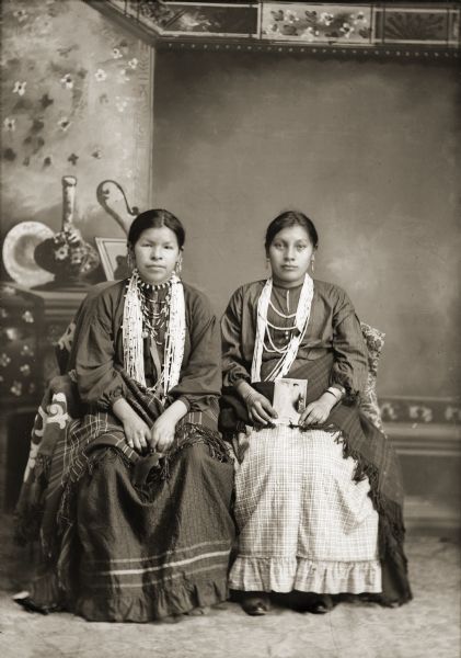 Studio portrait of two Ho-Chunk women, Frieda Blackcoon and Mary Cloud White (DaScotchAWinKah), posing sitting in front of a painted backdrop. They are both wearing traditional women's dresses (hinukwaje), several necklaces, file bracelets, rings, and earrings. Mary Cloud White is holding a Van Schaick cabinet card photograph on her lap.