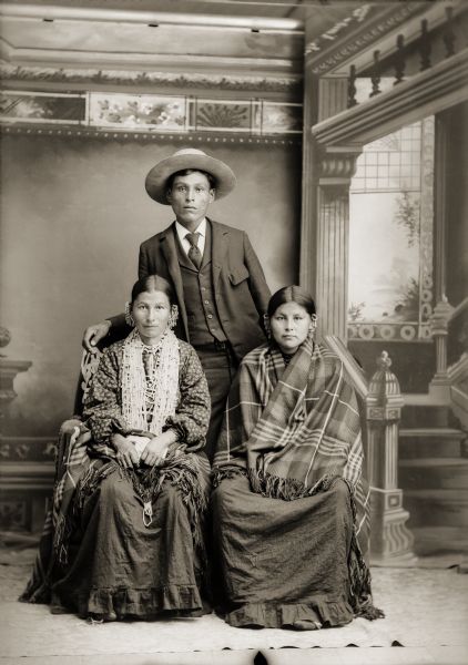 Studio portrait of Ho-Chunk group, from left to right, the wife of George (Lyons) Lowe (AhHaZheeKah), George Monegar, (EwaOnaGinaKah) and the sister of John Stacy (ChoNeKayHunKah), who are posing in front of a painted backdrop. The women are wearing several earrings, and John Stacey's sister, sitting on the left, is also wearing several necklaces and a shawl around her waist. George Lowe's wife, sitting on the right, has a shawl around her shoulders. George Monegar, standing behind the women, is wearing a suit, tie, and hat.