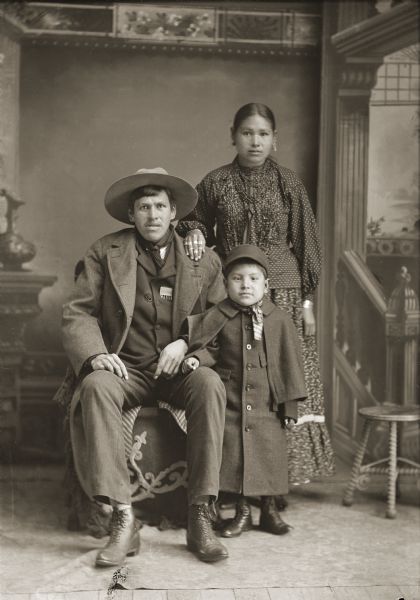 Full-length studio portrait of a family. (l to r) John Rogue (Coose He Rae Kaw) (son of Young Rogue [Wa Cho We Kaw] and [Wa Kon Hu Gi Win Kaw]) (from Winnebago, NE) sitting on the left wearing a heavy winter coat over a suit, with a bandana, and hat. His Ho-Chunk wife, Henukaw Davis, Rogue standing on the right with her right hand on the man's left shoulder, wearing a dark-colored blouse with light-colored polka dots, and several necklaces and earrings. Their son, Charles Rogue, stands with his right arm on the man's left leg, wearing a long overcoat and hat. In the background is a painted backdrop.