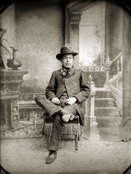 Studio portrait of John Johnson, a Nebraska Winnebago, posing sitting on a chair in front of a painted backdrop with his legs crossed. He is wearing a suit, a bandana around his neck, long earrings and a hat with a peacock feather in it.