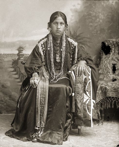Studio portrait of Ho-Chunk woman, Mabel White Blackhawk St. Cyr sitting in front of a painted backdrop with her left arm resting on a silk applique blanket. She is dressed in Ho-Chunk regalia, wearing several necklaces, earrings, rings, a traditional blouse, and stole.