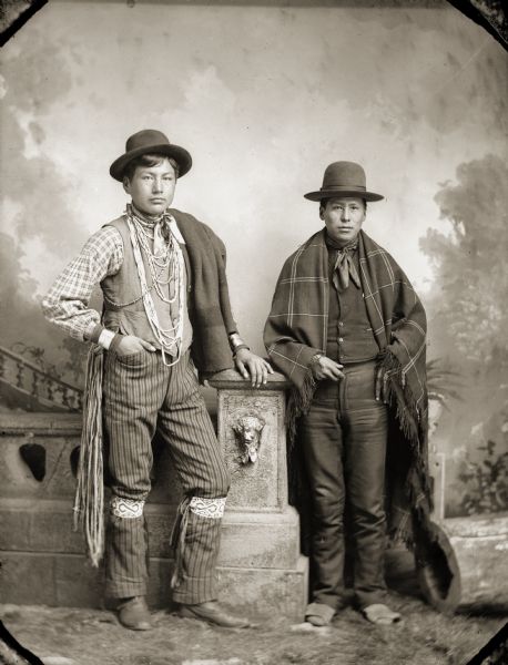 Full-length studio portrait of Ho-Chunk men, Frank Greencloud-Redcloud (NotchKeTaKah), left, and Ed Greengrass (CheWinCheKayRayHeKah). Frank Greencloud-Redcloud is wearing beaded-garters and has a Hudson's Bay wool blanket on his shoulder. He also is wearing a vest, a hat, several necklaces, striped pants, and file bracelets. On the right, Ed Greengrass is wearing a vest, bandana, hat, and shawl over both shoulders. Both men are posing in front of a painted backdrop and are leaning on a prop stone wall.