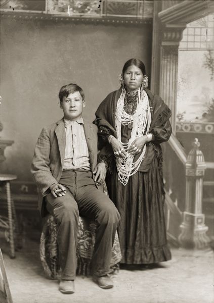Full-length studio portrait in front of a painted backdrop of Frank Browneagle (HeWaKaKayReKah), a Ho-Chunk man, and Annie Snake (KhaWinKeeSinchHayWinKah) posing in front of a painted backdrop. On the left, Frank Browneagle is sitting, wearing a suit coat, and holding a cigar in his right hand. On the right, Annie Snake is standing and wearing a shawl around her shoulders, and jewelry including earrings, necklaces, rings, and file bracelets. Snake was a member of the Nebraska Winnebago.