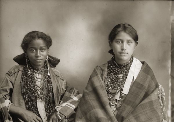 Waist-up studio portrait of two young Ho-Chunk women posing sitting, wearing beaded necklaces and long earrings, and wrapped in shawls. Carrie Elk (ENooKah), left, daughter of Lucie Elk, is of mixed African-American and Ho-Chunk heritage, though she identified herself as Ho-Chunk. Annie Lowe Lincoln (Red Bird), right, is the daughter of King of Thunder. Annie and Carrie were cousins.