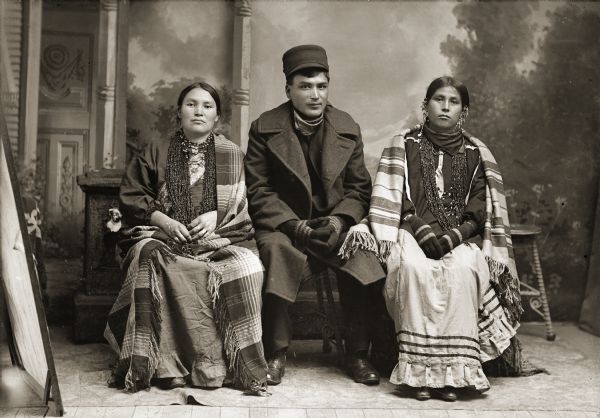 Full-length studio portrait of a Ho-Chunk group, Susie Decorra Thundercloud (UkSeKaHoNoKah), left, Albert (Robert) Henry (HeKeMeGa), center, and Dora Monegar Wallace Green (Snake) (ChePinChayWinKah), right. The women are wearing shawls, earrings, and bead necklaces. They are sitting on either side of Albert Henry who is wearing a coat, hat, and mittens. They are posing in front of a prop stone wall and painted backdrop.