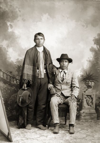 Full-length studio portrait of two Ho-Chunk men, Charles (Charlie) Sine Jr., sitting, and George Greencrow, standing, in front of a painted backdrop. George Greencrow, on the left, is wearing moccasins and a shirt with two long ribbons hanging down the front. The ribbons have small silver brooches (hiiwapox) attached. Charles Sine Jr. is sitting and wearing a suit jacket, trousers, and a hat.
