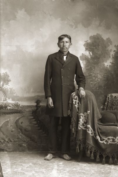 Full-length studio portrait in front of a painted backdrop of a Ho-Chunk man, James (Jim) Sine (KahHeHoNoNeeKah) wearing a long coat, tie, trousers, and beaded moccasins. He is standing next to a stuffed chair covered with a fringed blanket, with a hat on the seat.