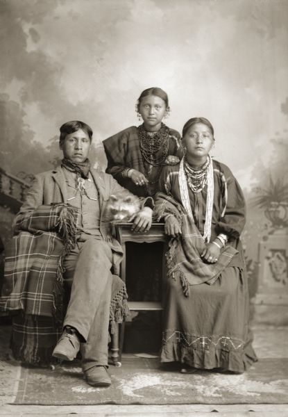 Full-length studio portrait of a Ho-Chunk group in front of a painted backdrop. On the left is William Massey (ChawRoChooChayKah), sitting with his arm around his pet raccoon and, Grace Decorra Twocrow Winneshiek Massey (MauHeHutTaWinKah), sitting on the right. Standing behind them in the center is Fanny C. Whitedog (HoChunTaWinKah). The women are wearing some regalia including necklaces and earrings, and William Massey is wearing a suit and holding a plaid shawl.