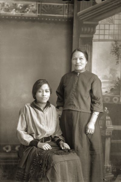 Studio portrait in front of a painted backdrop of two Ho-Chunk women, Carrie Elk (ENooKah) posing sitting with a fringed shawl on her lap, and an unidentified woman wearing a Christian cross, who is standing. Both women are wearing narrow choker necklaces and contemporary clothing.